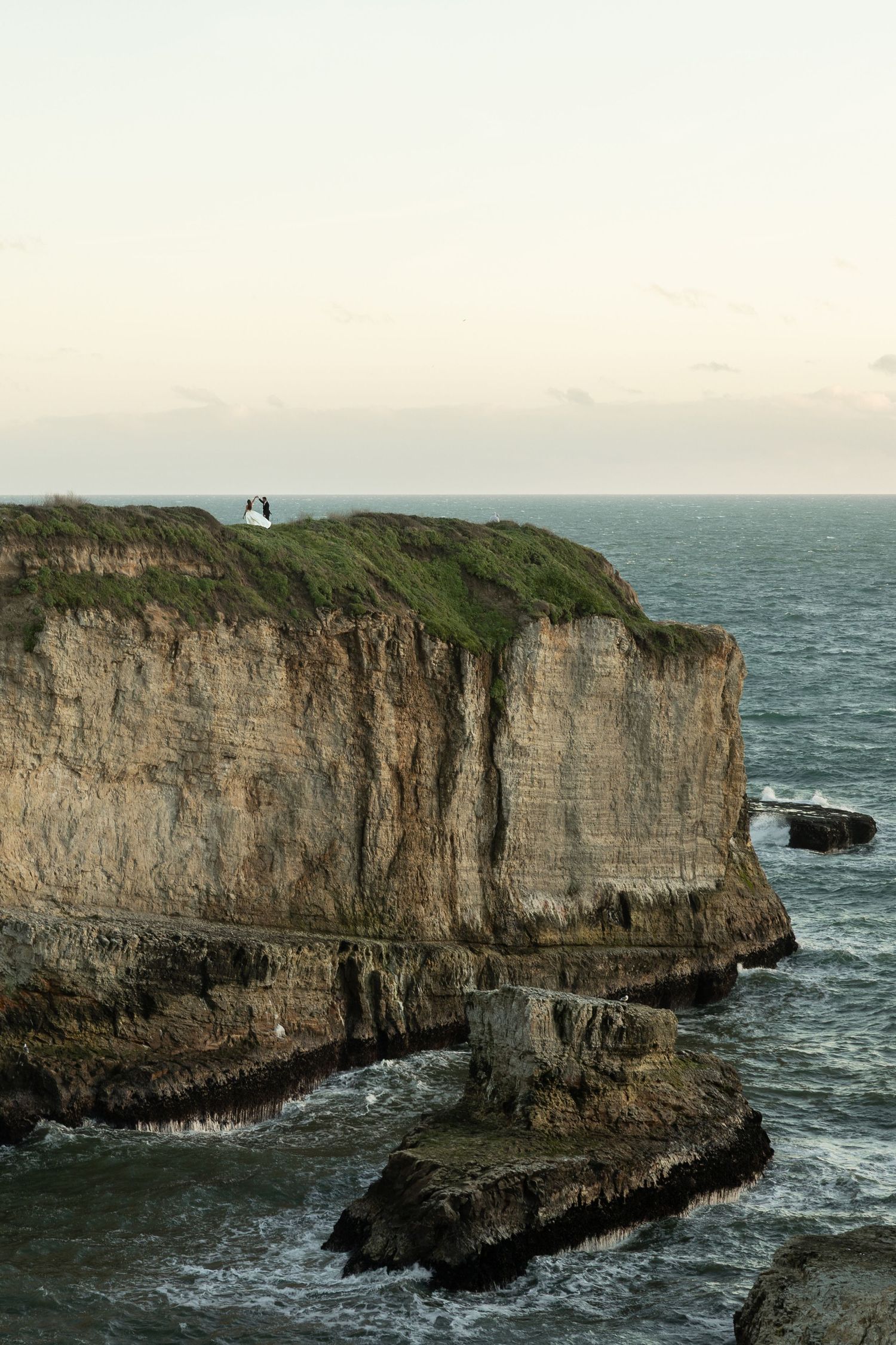 Couple standing on dramatic ocean cliffs edge with turquoise sea views and rocky coastline below at dusk at Shark Fin Cove.