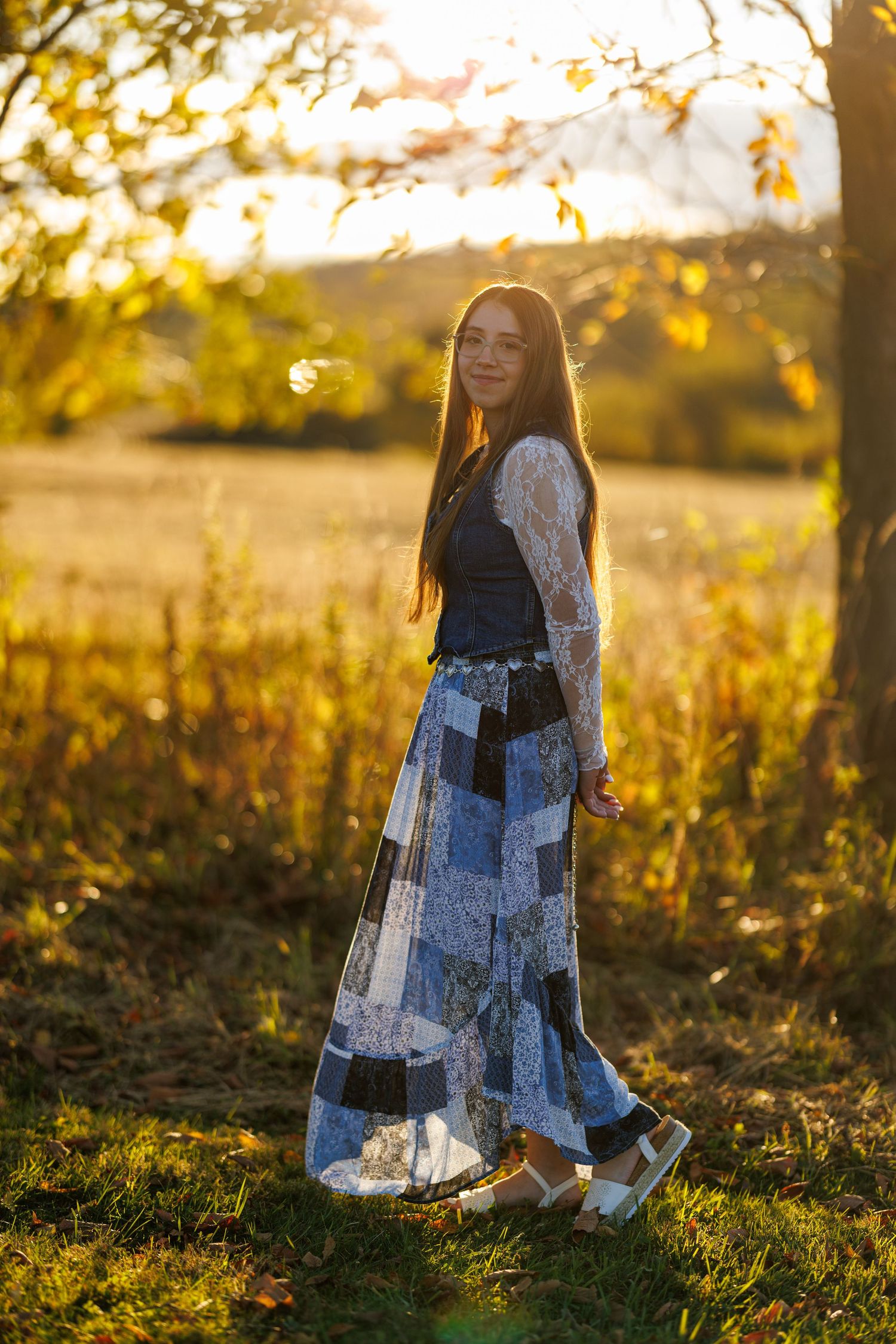 Teenage girl in patchwork blue maxi skirt and lace top posing in golden autumn sunset light outdoors.