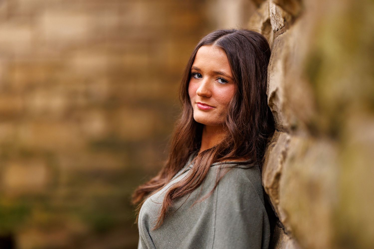 Young woman with long brown hair leaning against stone wall in olive green sweater.