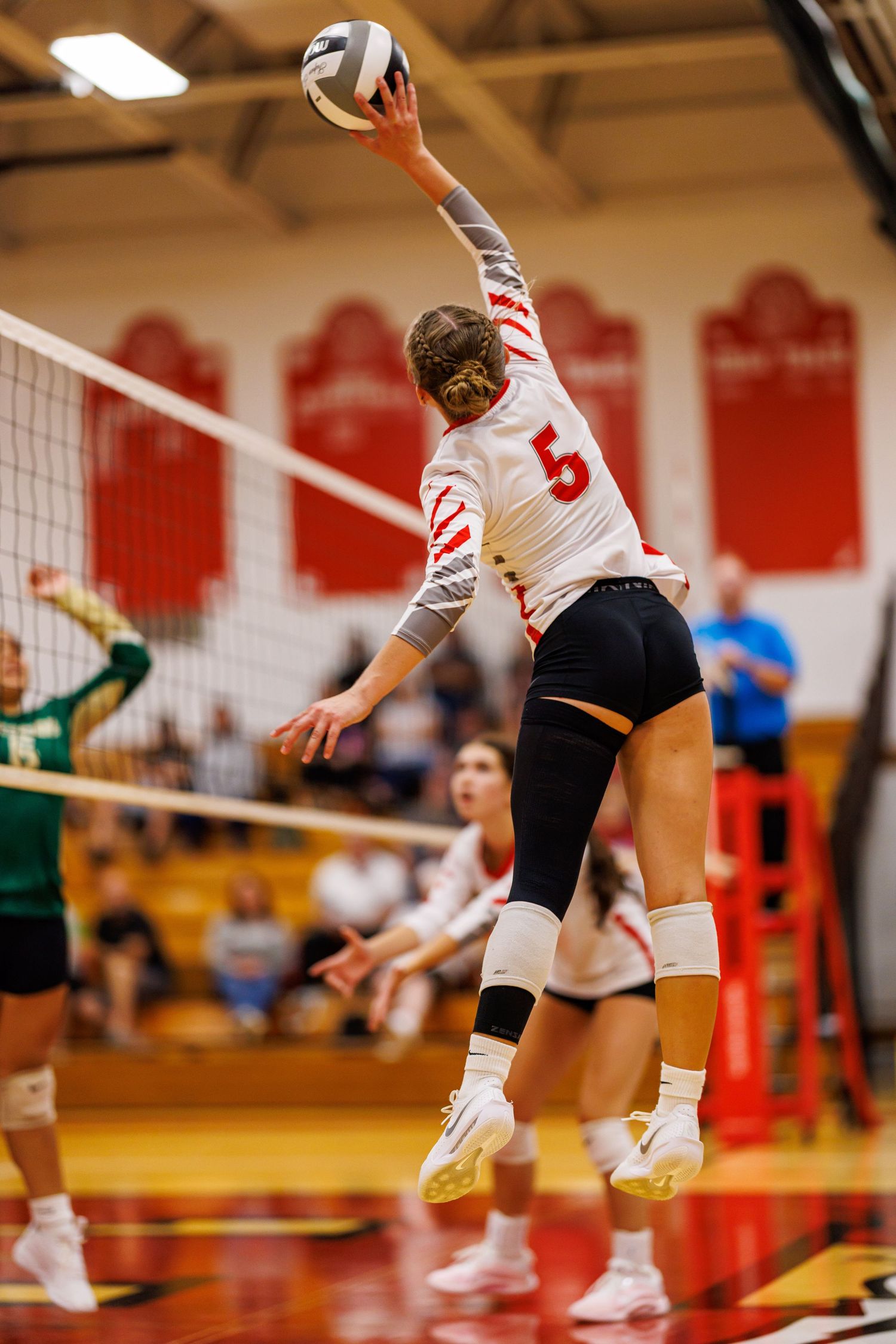 Female volleyball player wearing number 5 jersey jumping to spike the ball during an indoor high school match.