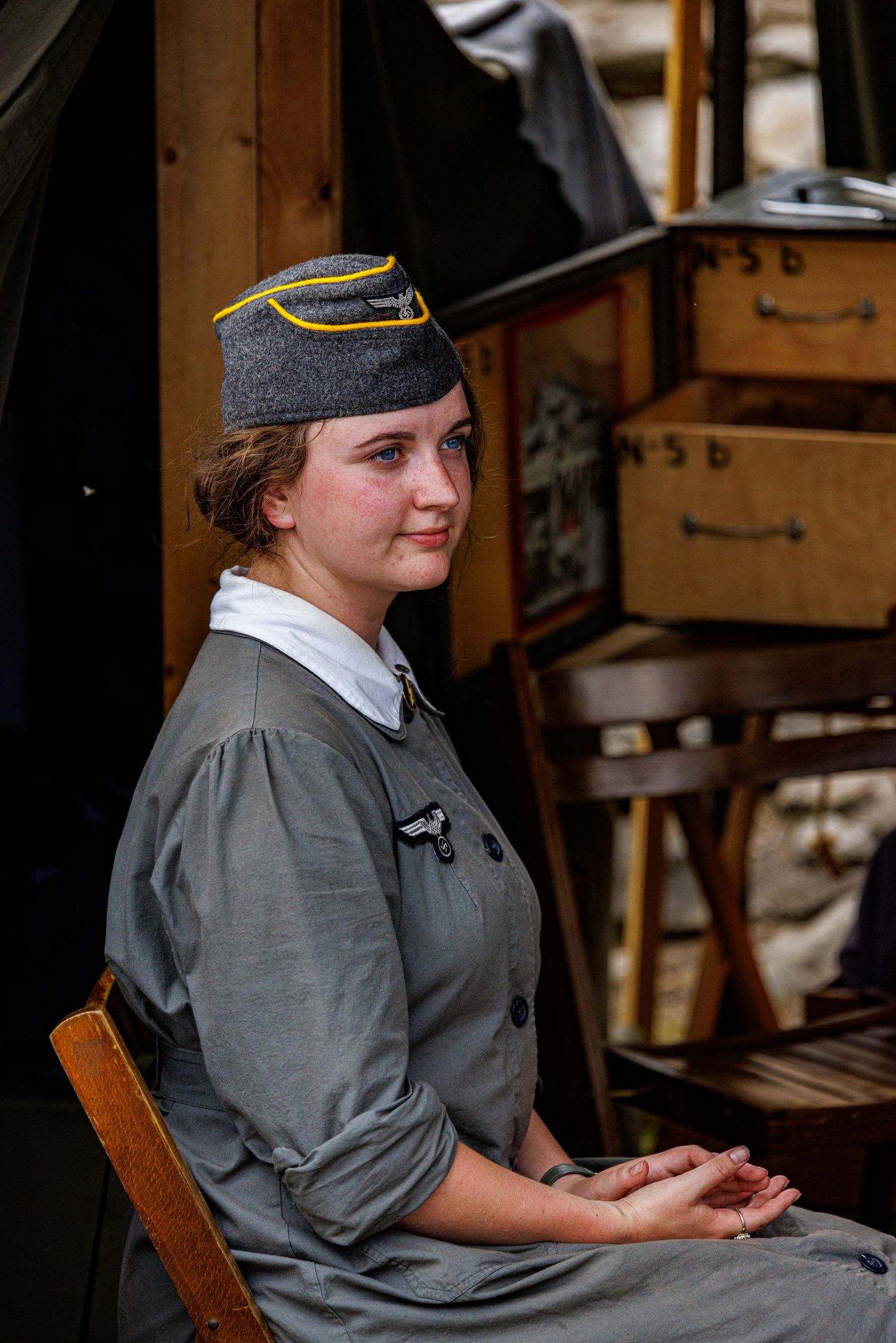 Young woman in WWII German uniform with garrison cap seated at historical military reenactment.