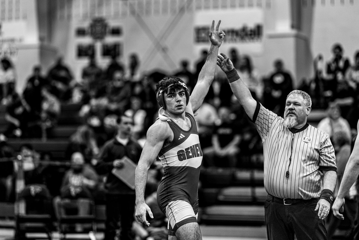 A referee raises a young wrestler's hand in victory after a wrestling match in a packed gymnasium.