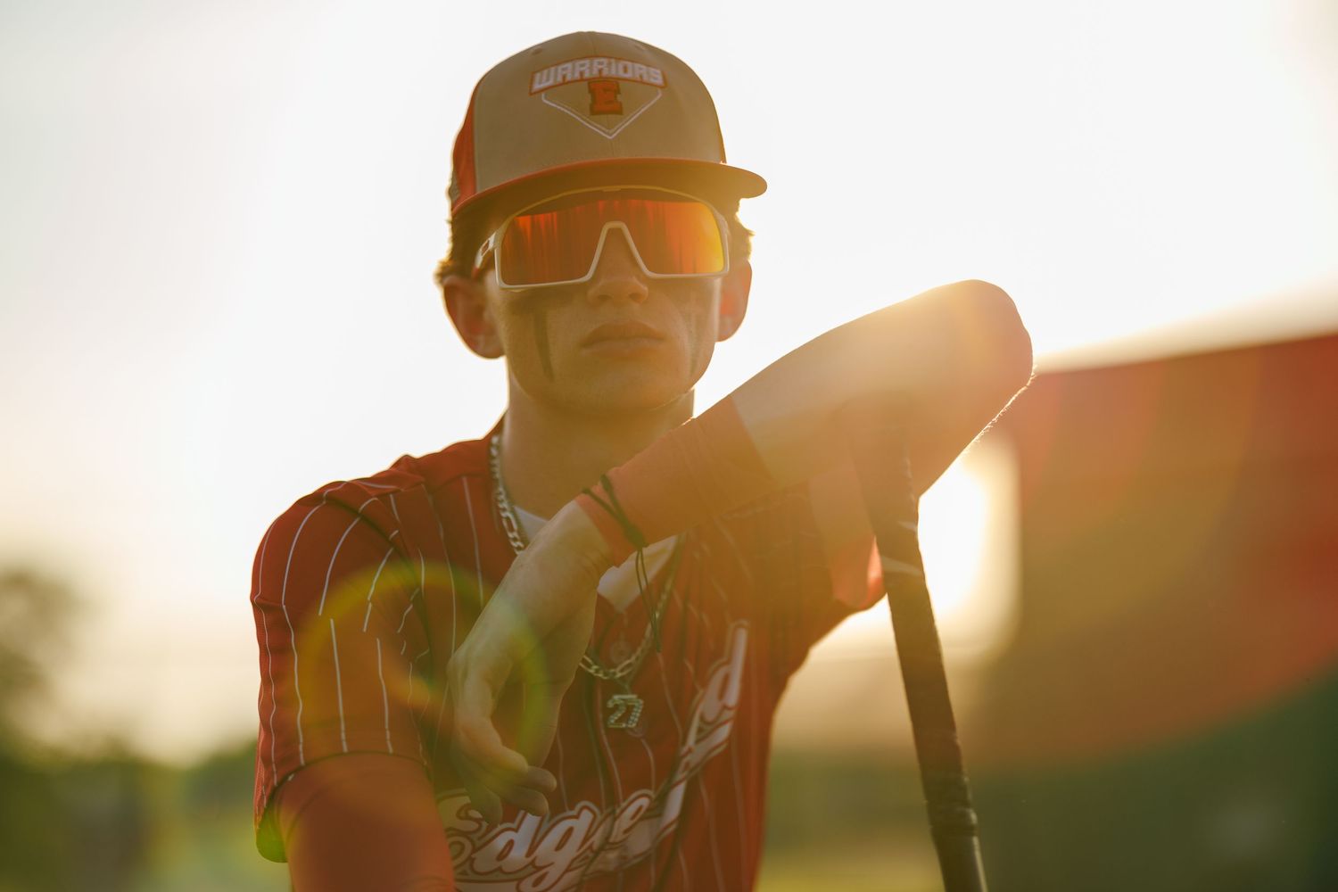 Young baseball player in orange jersey and sunglasses poses with bat in golden sunset light.