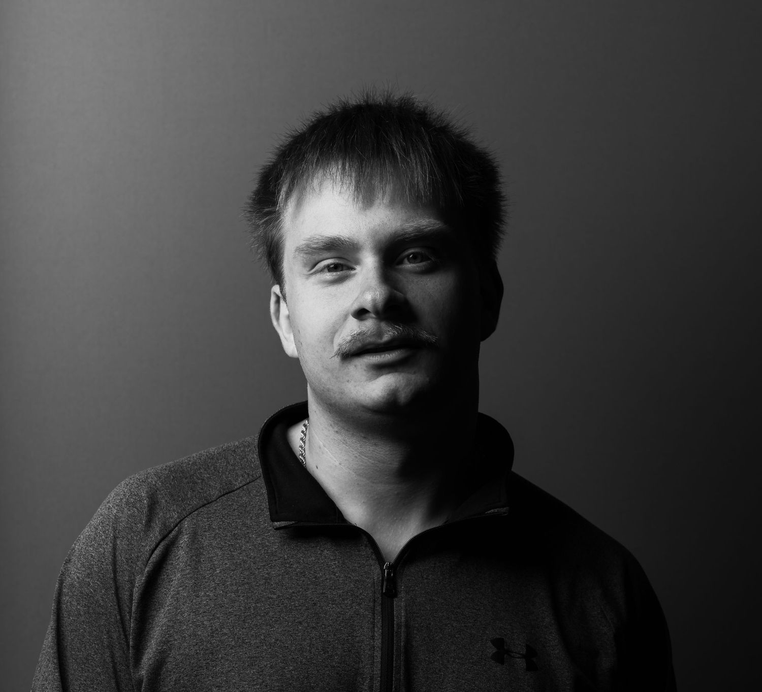 Black and white portrait of a young man in a gray Under Armour jacket against a dark background.