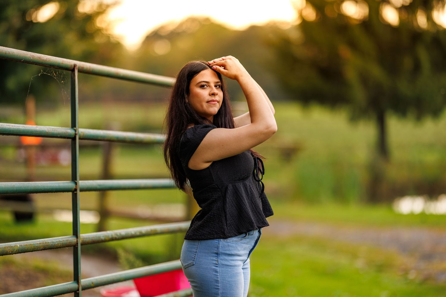 Brunette woman posing by metal fence in black top and jeans at sunset.