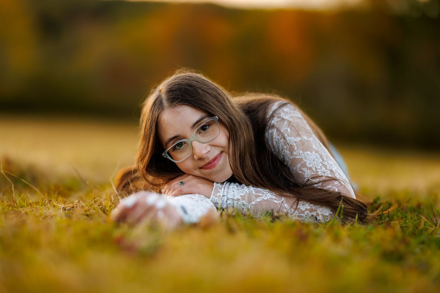 Smiling teenage girl with glasses lying on grass in autumn field with warm bokeh background.