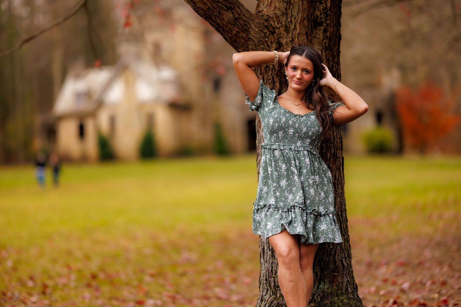 Teen girl in floral green dress posing against tree with historic stone building backdrop.