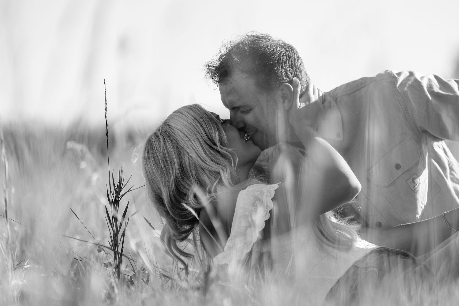 A couple shares a romantic kiss in a tall grass field in a black and white photo.