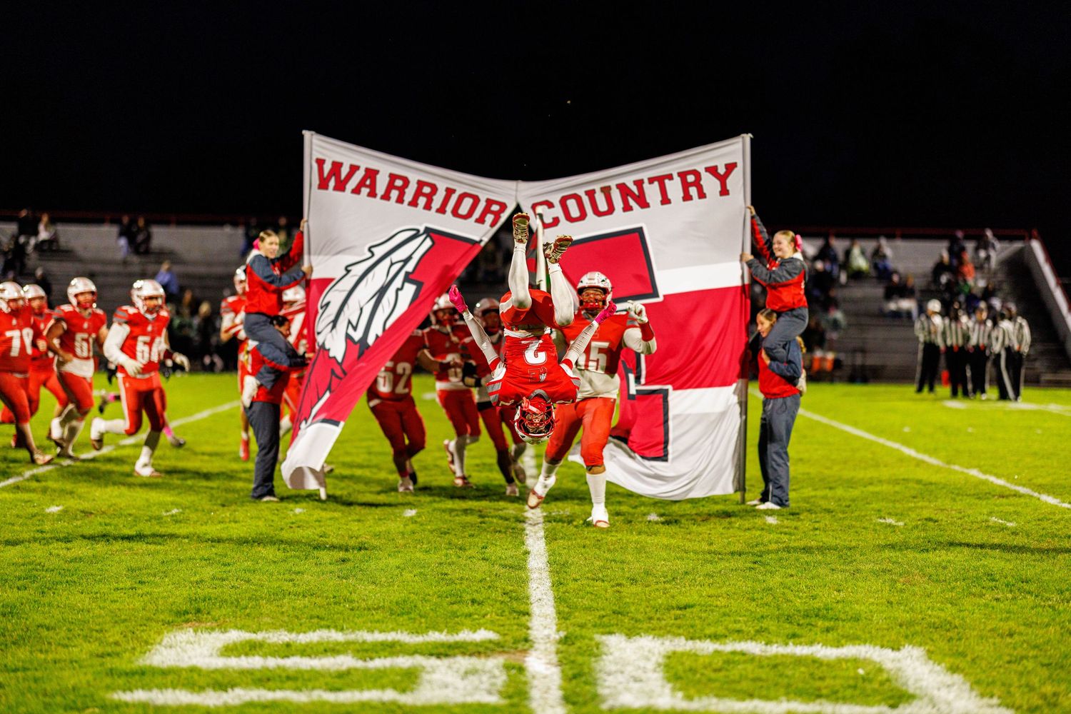 High school football players burst through Warrior Country banner under Friday night lights during exciting game entrance.