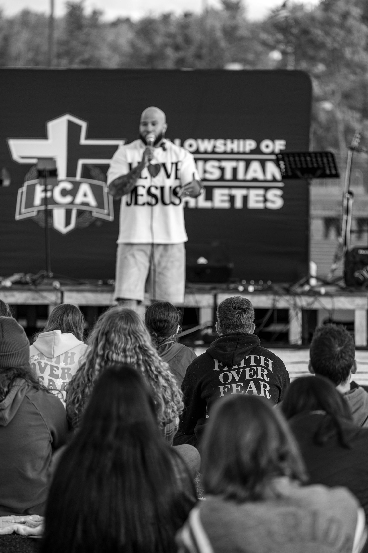 Speaker in I Love Jesus shirt addresses crowd at Fellowship of Christian Athletes outdoor event.