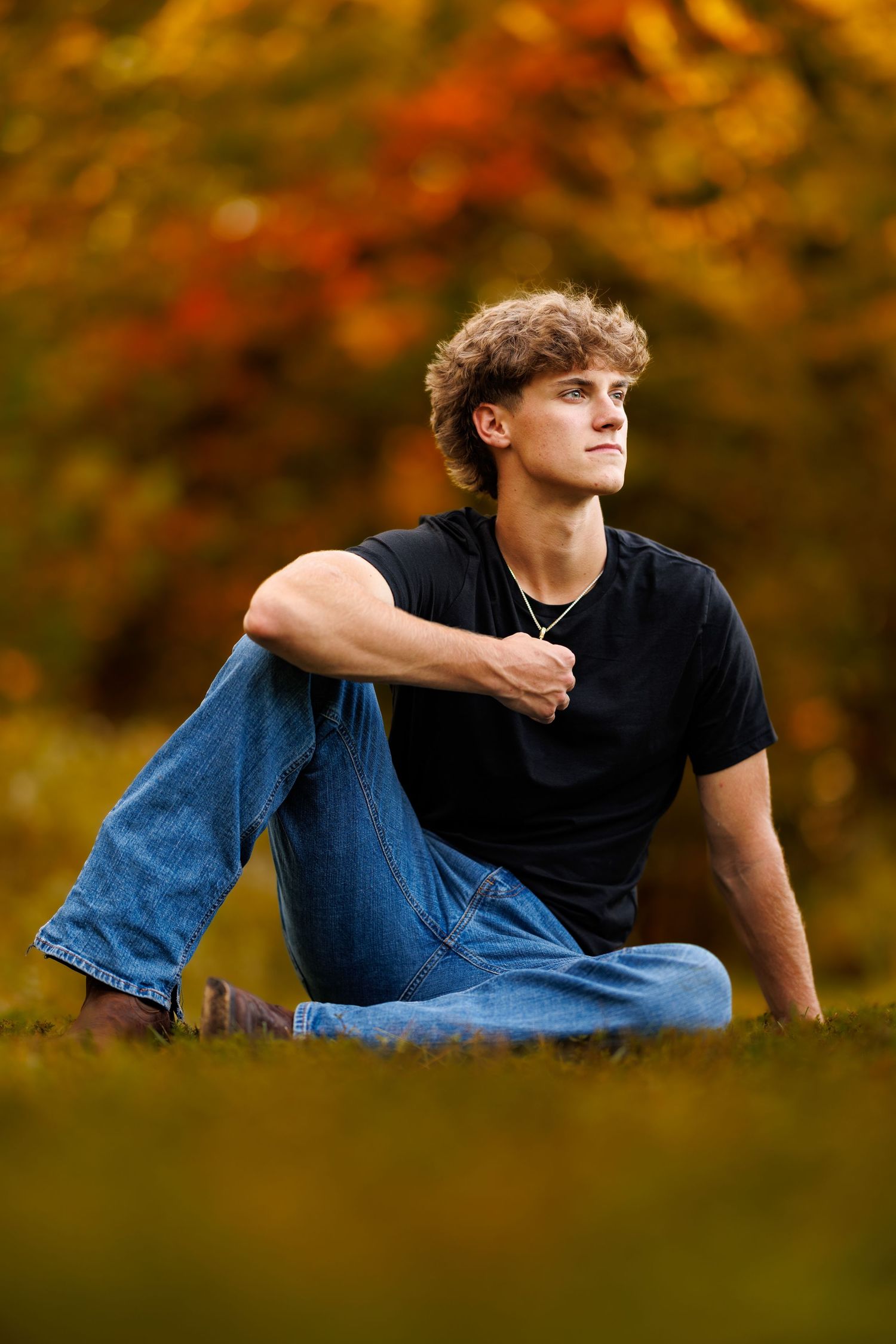 Teen boy in black shirt and jeans sitting on grass with vibrant fall foliage background.
