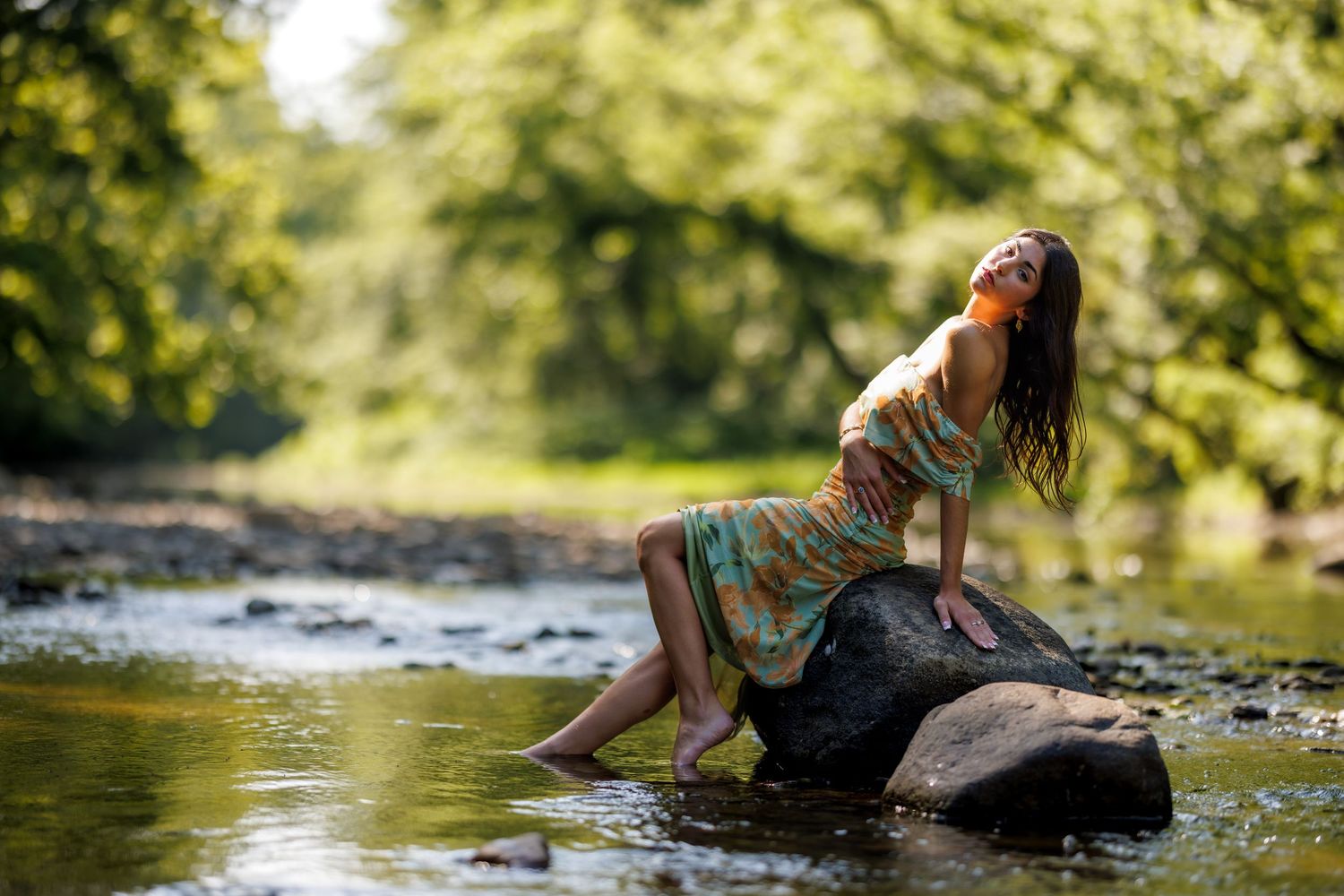 A woman in a floral dress sits on a rock in a shallow forest stream.