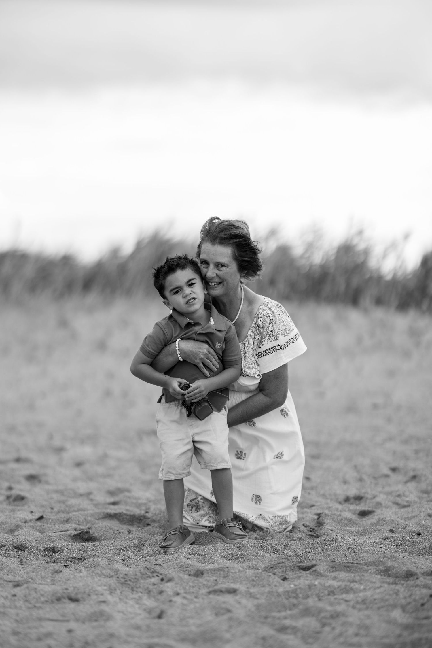 Black and white photo of a grandmother hugging her young grandson on a sandy beach.