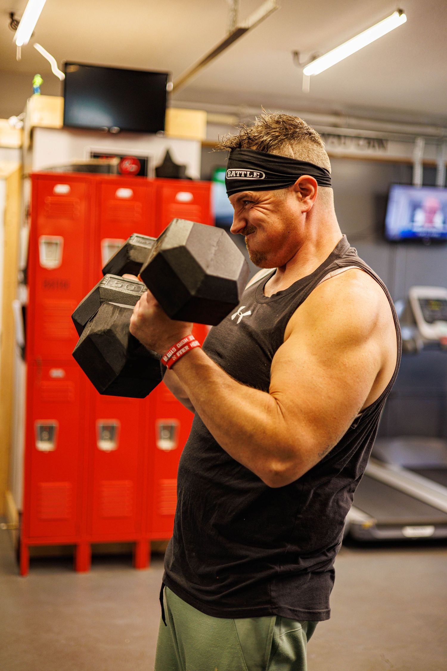 Muscular man in black tank top curls dumbbell while wearing headband in gym with red lockers.