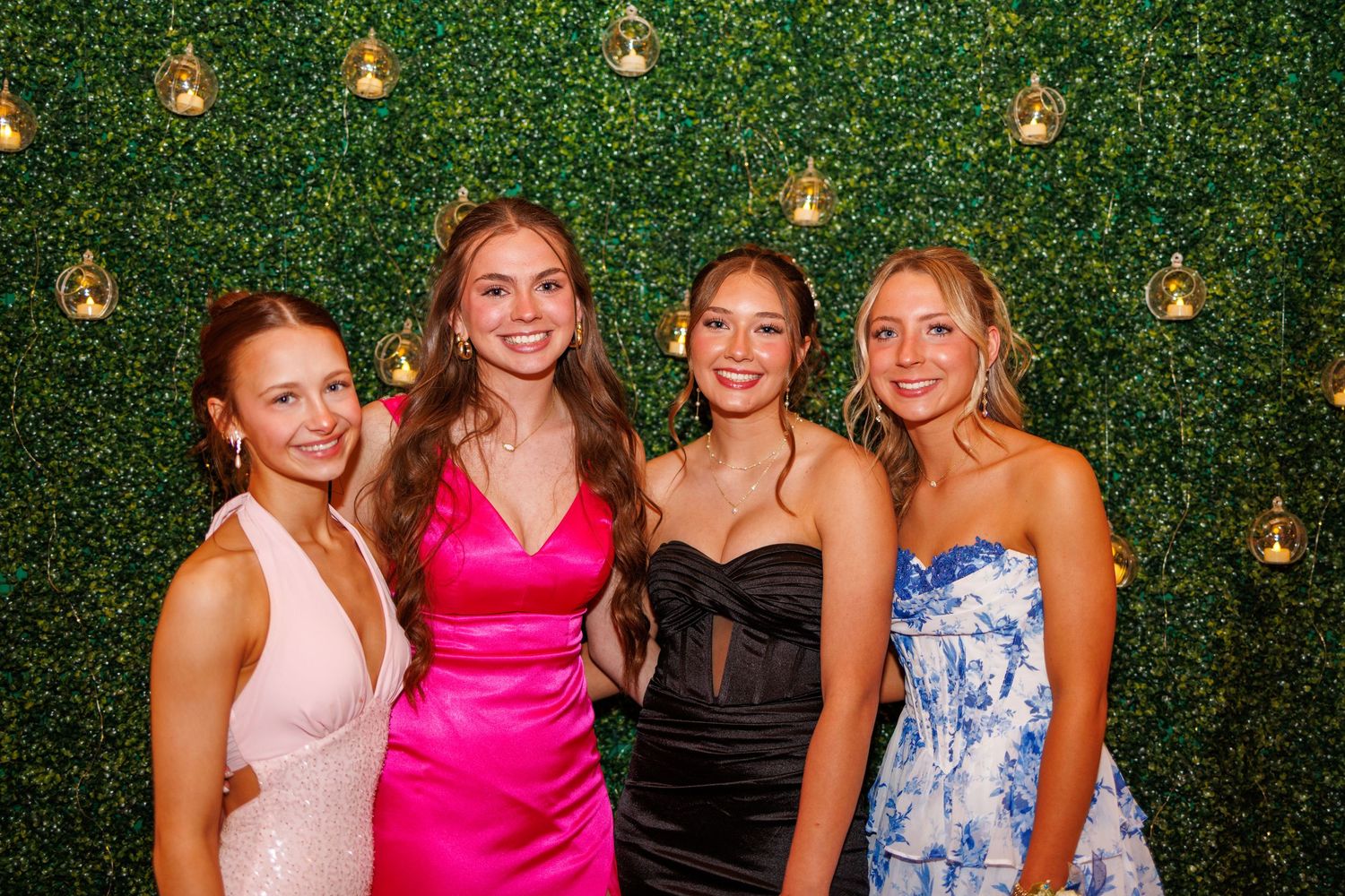 Four young women in elegant prom dresses posing together in front of a green hedge photo backdrop.