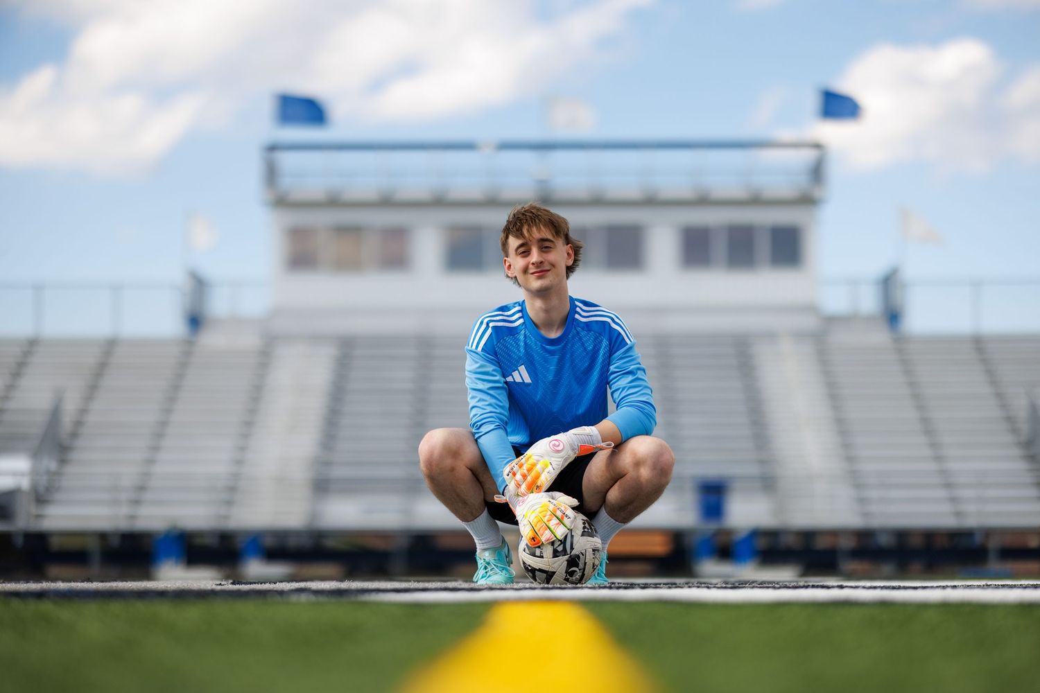 Teen soccer player crouching on field holding ball in front of stadium bleachers on a cloudy day.