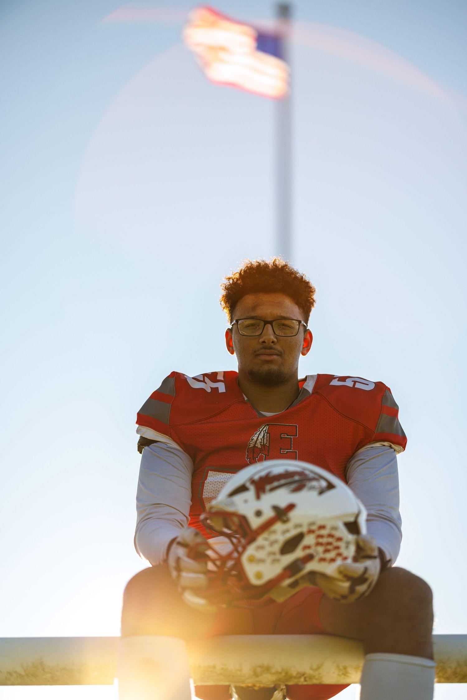 Football player in red uniform holding helmet seated before American flag in dramatic backlit sky.
