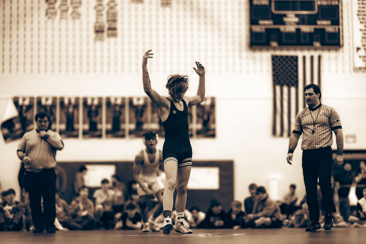 A young wrestler celebrates victory with arms raised on the mat in a vintage-toned gymnasium photo.