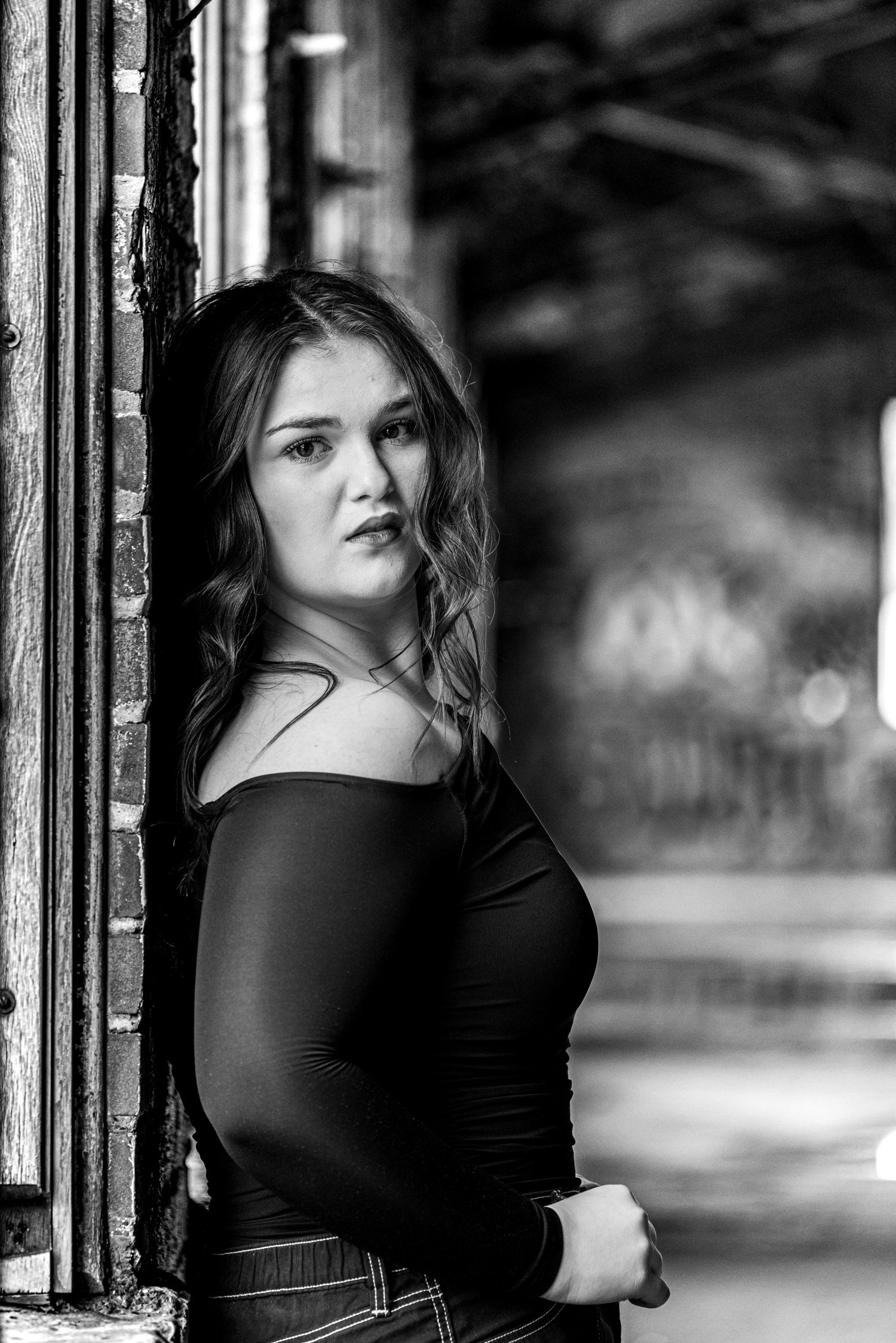 Black and white portrait of young woman in off-shoulder top leaning against brick wall in urban setting.