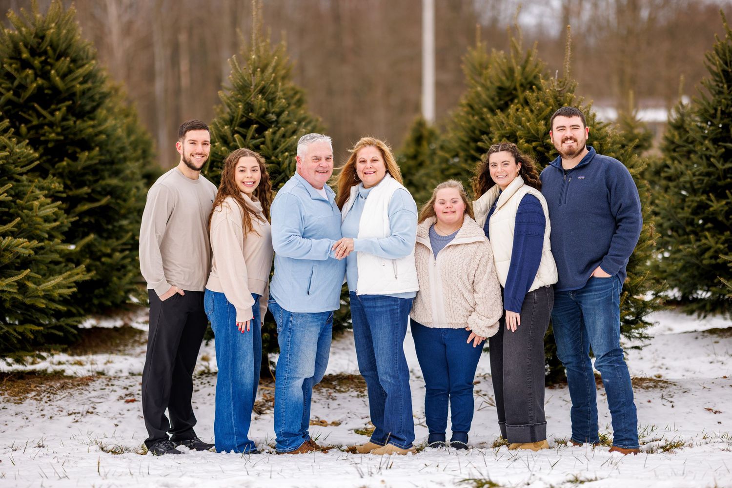 A family of seven poses together smiling in a snowy Christmas tree farm during winter.