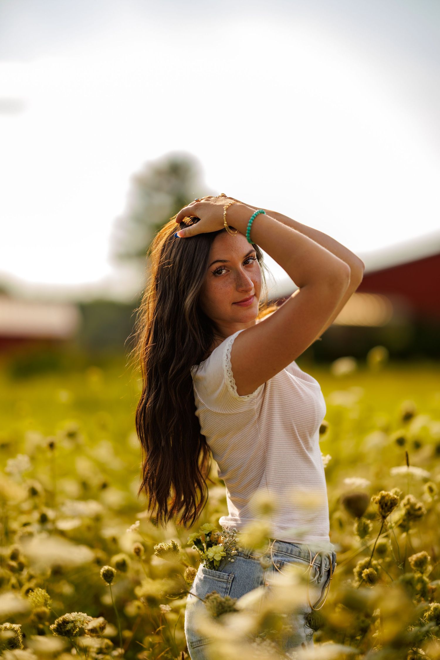 Young woman in white t-shirt and jeans standing in a green field of wildflowers at golden hour.