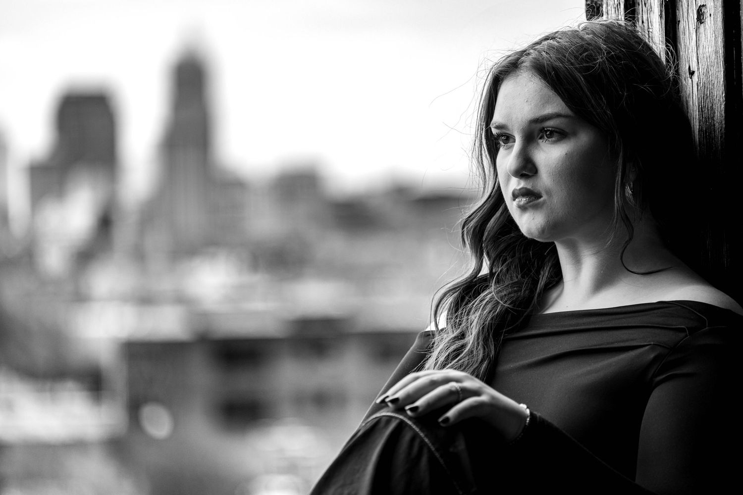 Black and white portrait of young woman with wavy hair gazing thoughtfully with city skyline behind her.