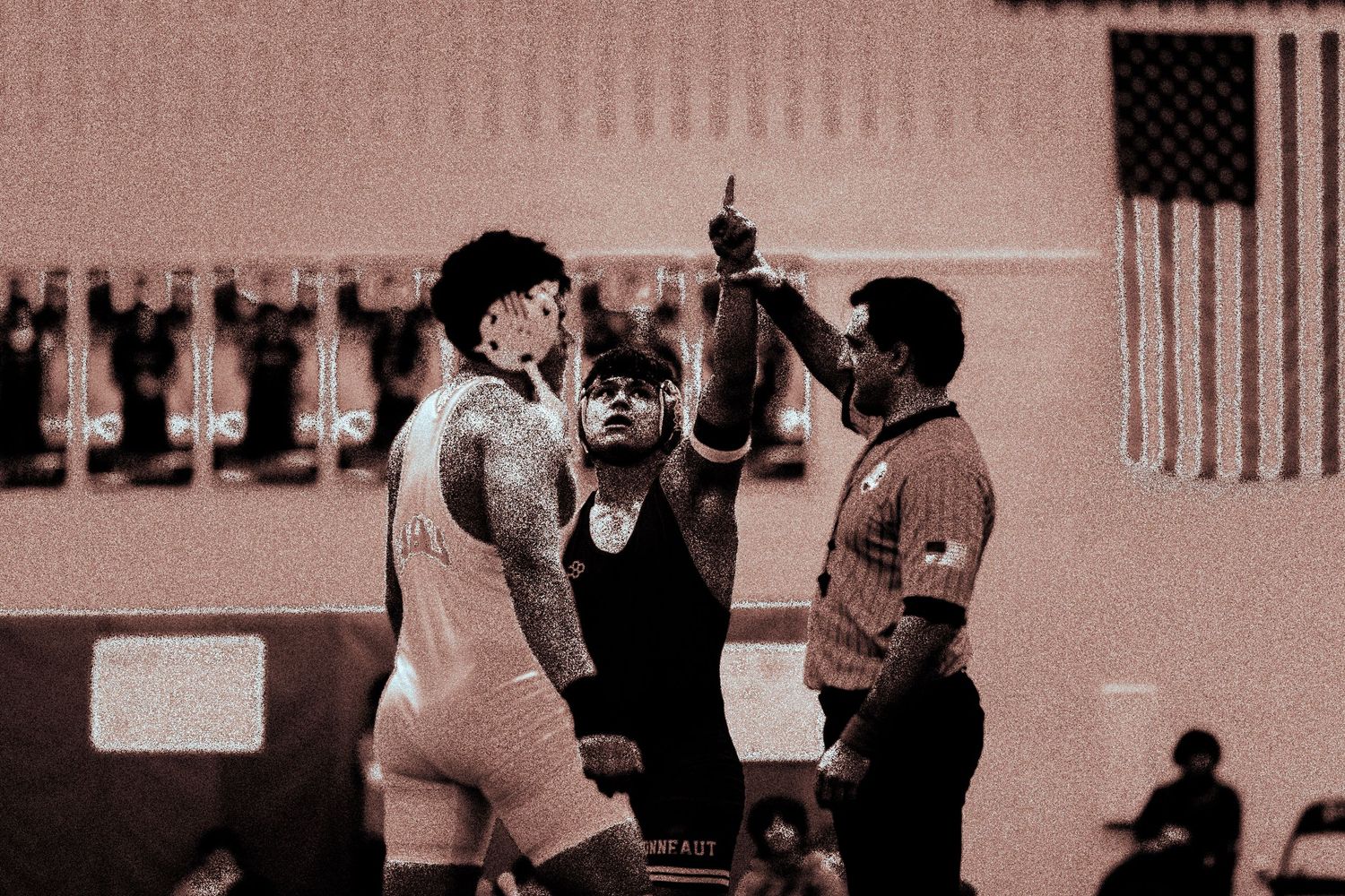 A referee raises a wrestler's hand in victory after a competitive wrestling match in a gymnasium.