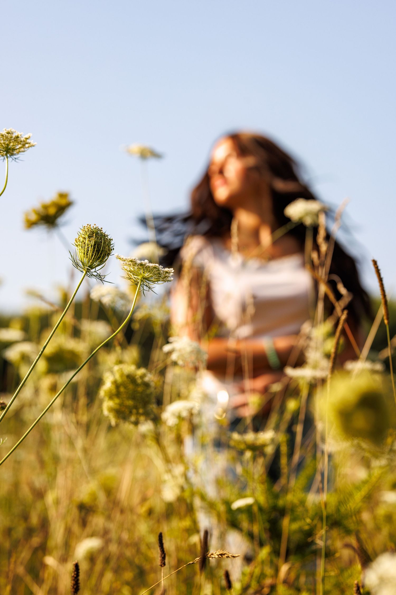 Blurred young woman standing in a sunny field with white wildflowers in sharp focus in the foreground.