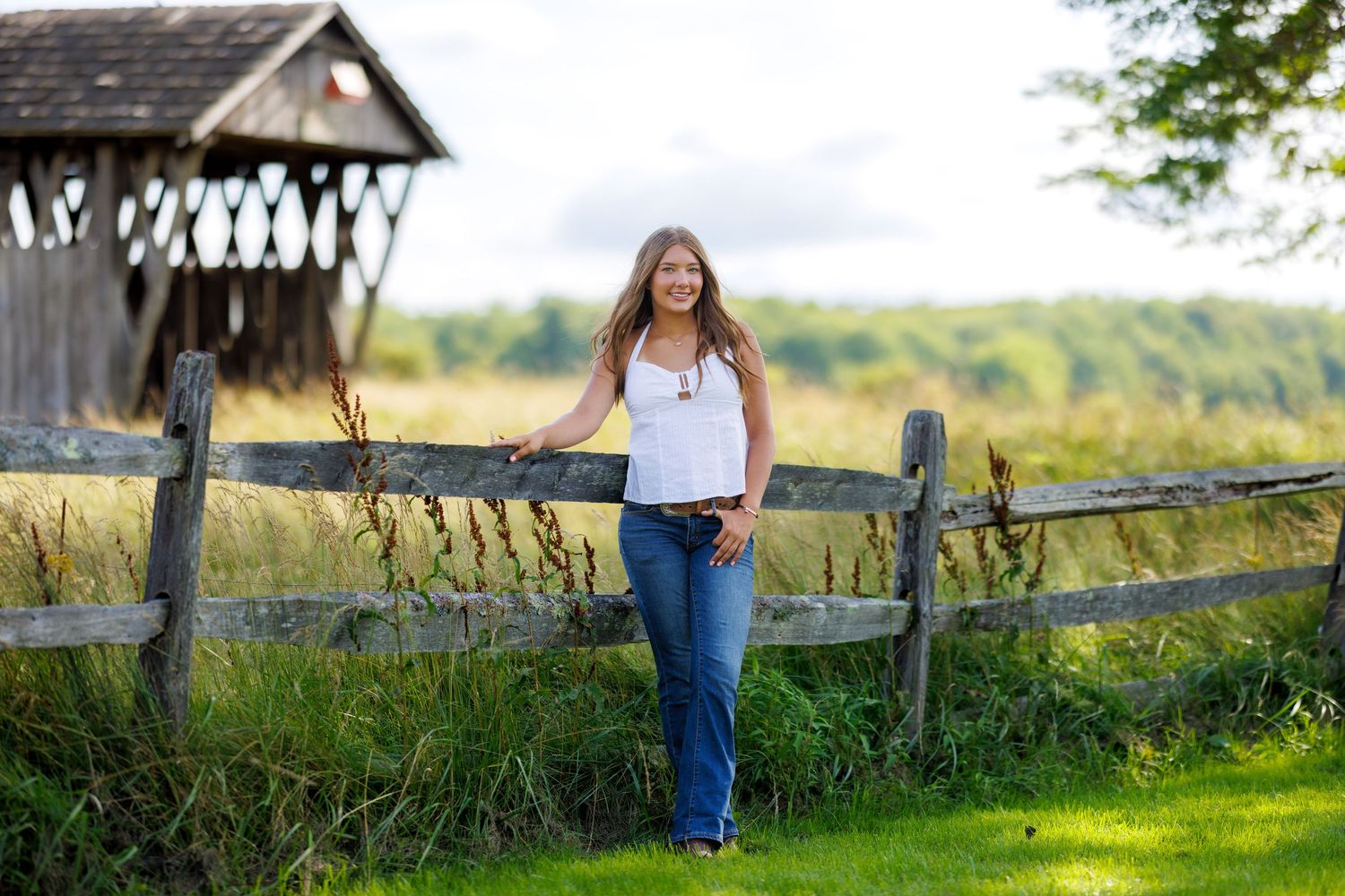 A teenage girl in jeans and white top leans on a wooden fence near a covered bridge.
