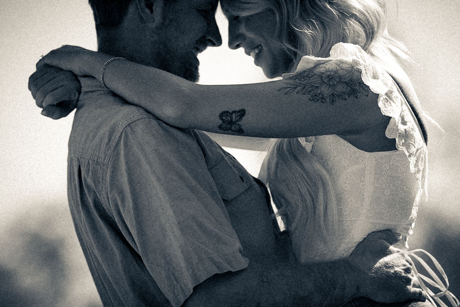 Intimate black and white portrait of couple holding each other close outdoors, butterfly tattoo visible.
