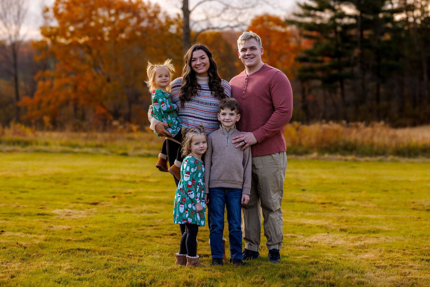 Family of five smiling together in a field with colorful autumn foliage in background.