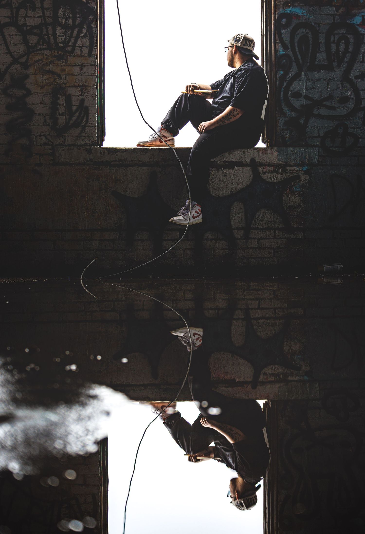 Man sitting in graffiti-covered abandoned building window with fishing rod reflected in still water below.