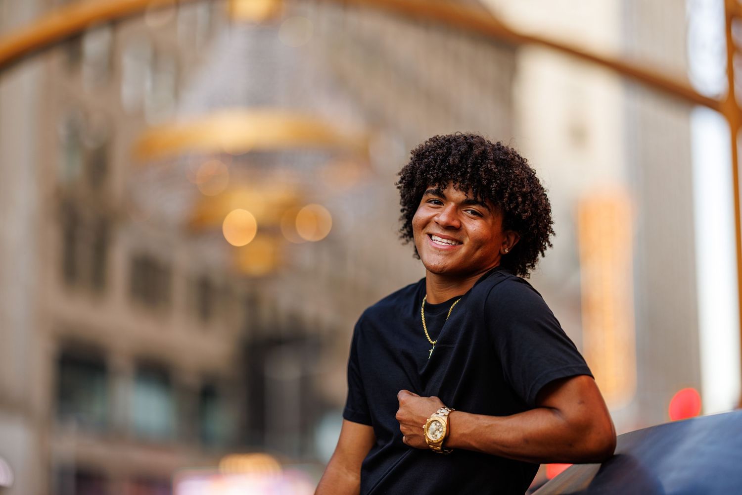 Smiling young man with curly hair wearing gold chain seated outdoors in urban setting.