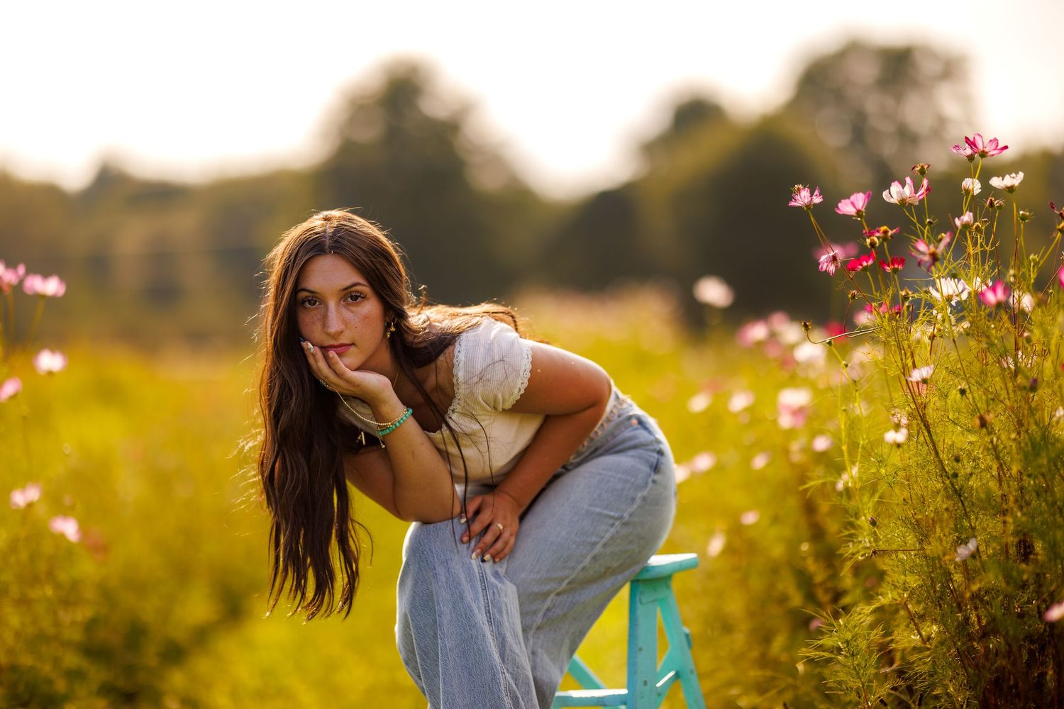Young brunette woman leaning on a teal stool surrounded by pink wildflowers in a golden meadow.