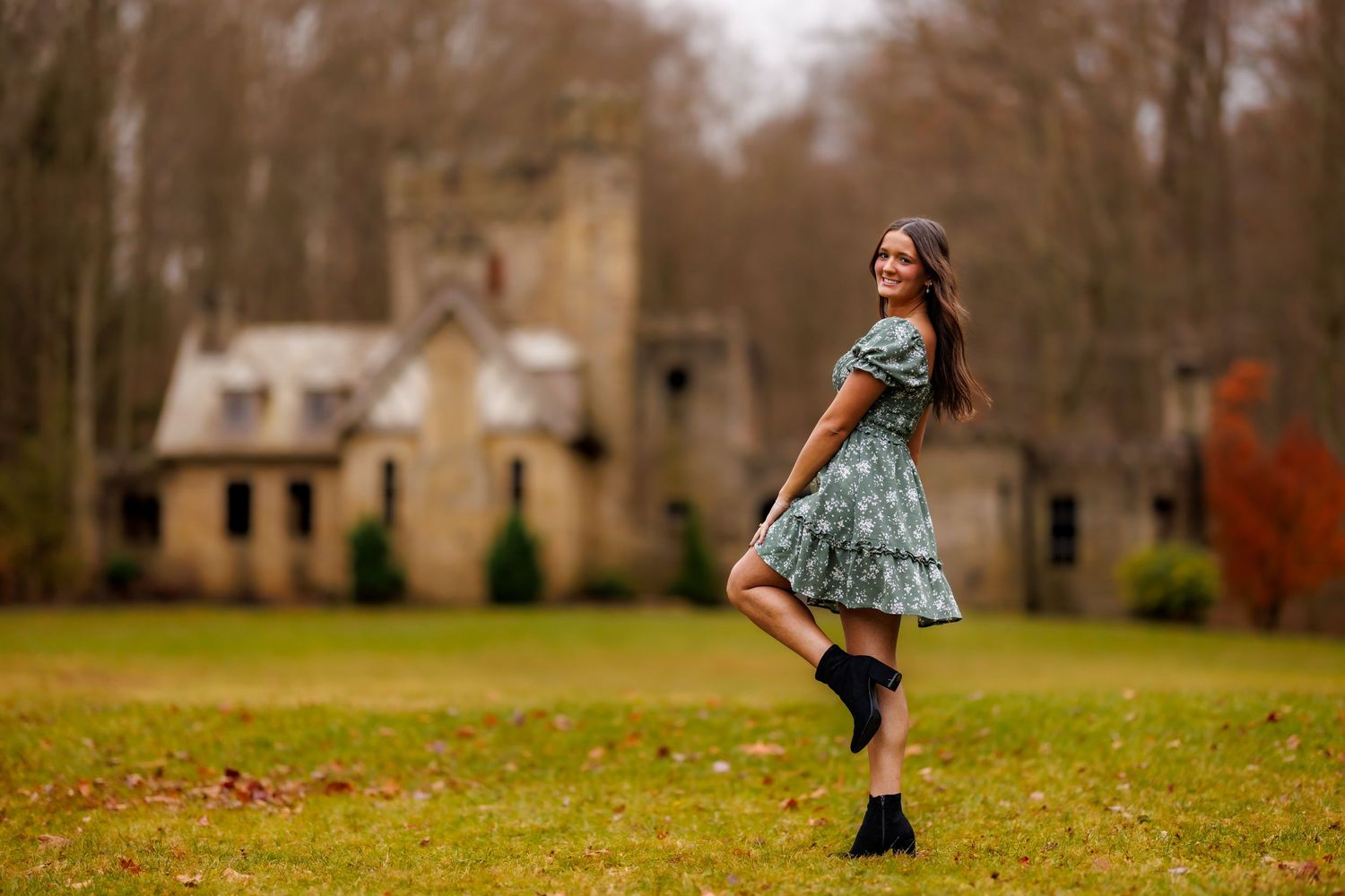 Teen girl in floral green dress spinning joyfully on lawn near rustic stone building.