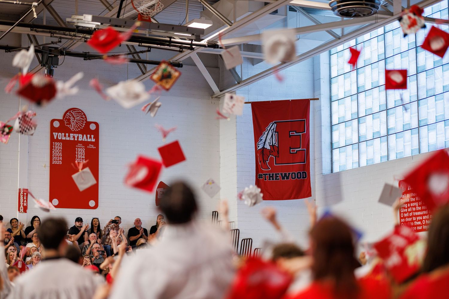 Red and white graduation caps flying in the air during a high school commencement ceremony in a gymnasium.