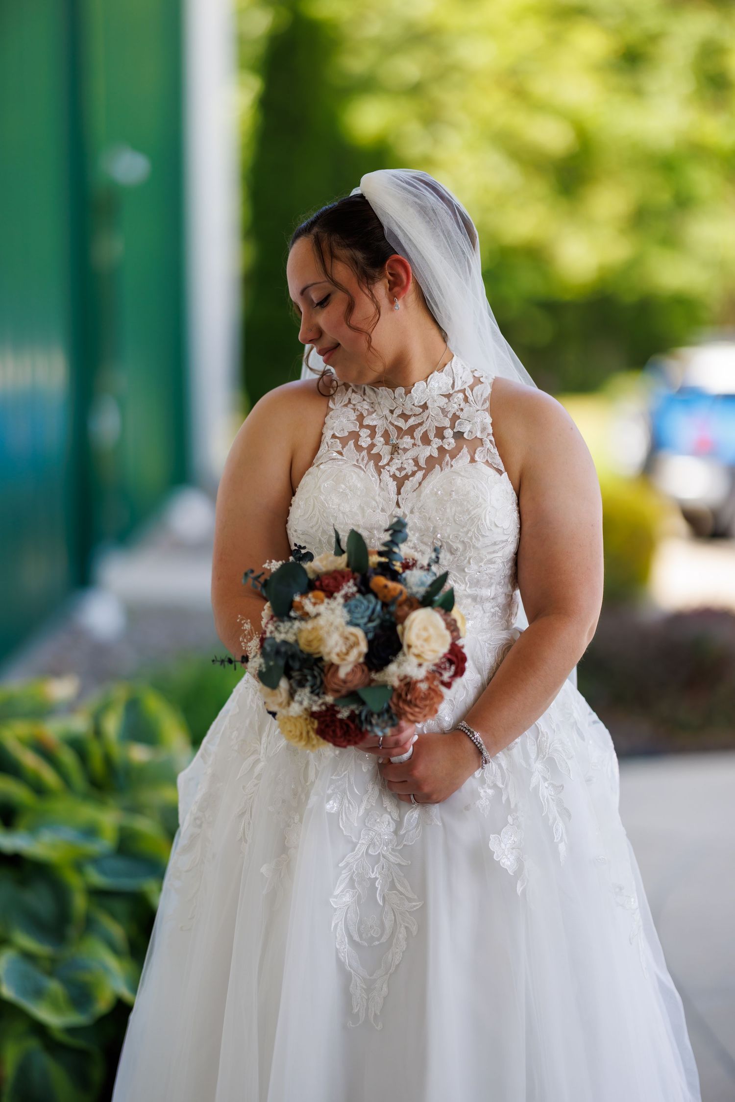 Bride in lace halter gown holding colorful bouquet with veil outdoors.