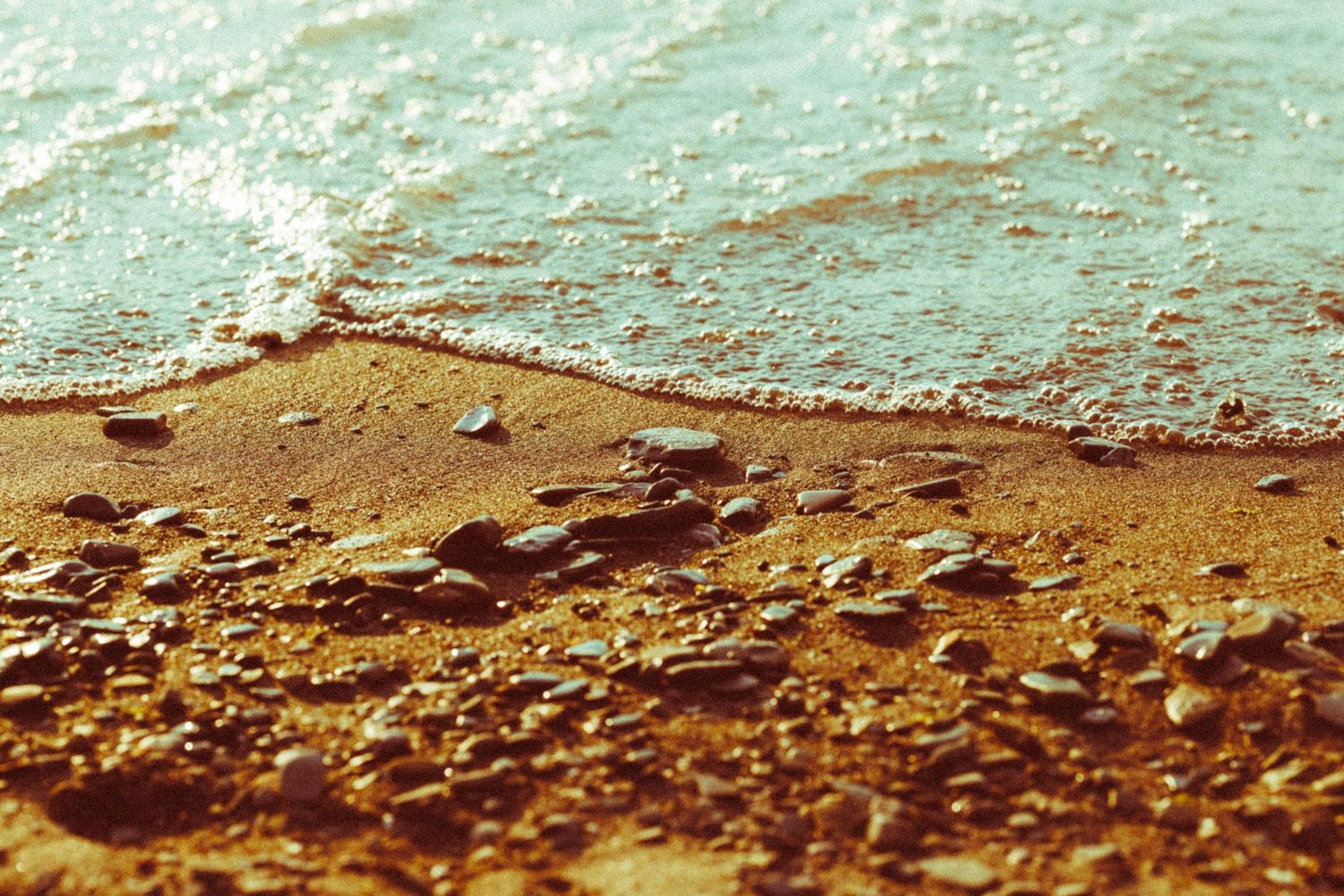 Close-up of golden sandy beach with small pebbles and foamy ocean waves washing ashore in warm tones.