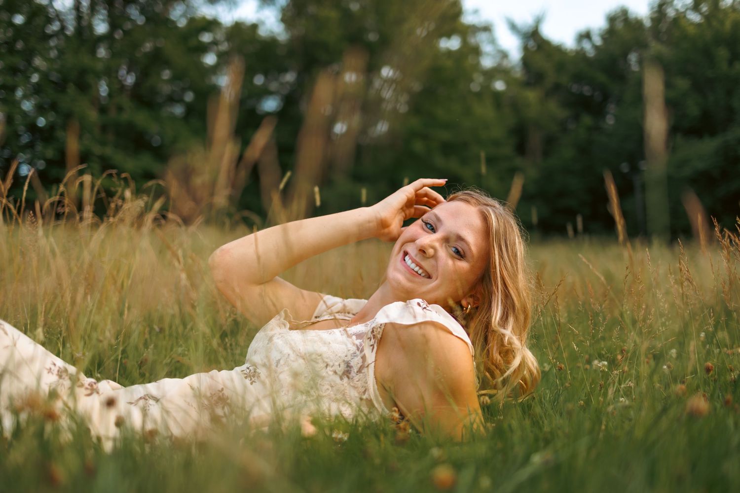 Smiling blonde woman in a white lace dress lying in a sunny green meadow with trees in background.