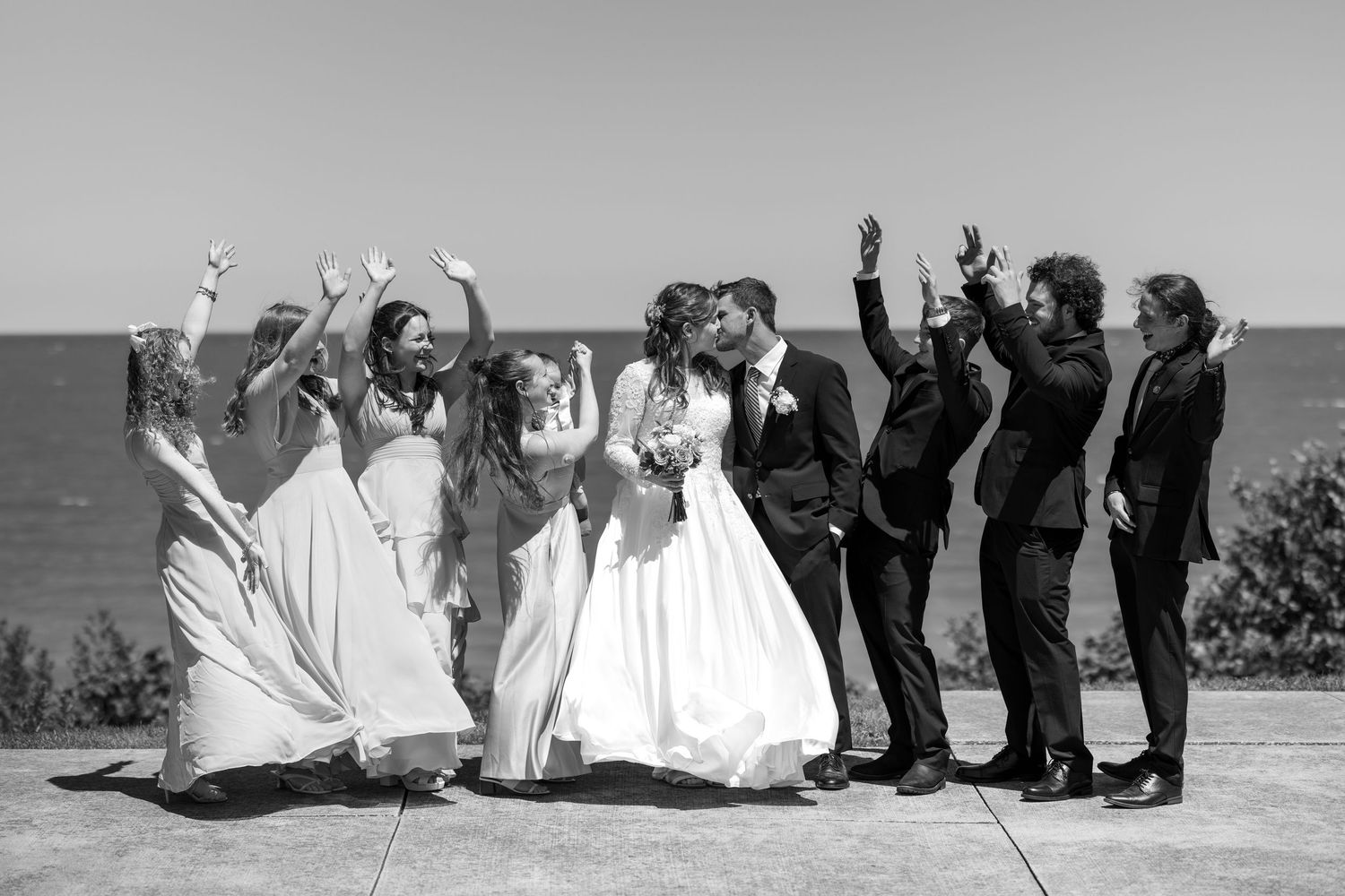 Wedding party celebrating with raised hands while bride and groom kiss near waterfront in black and white.
