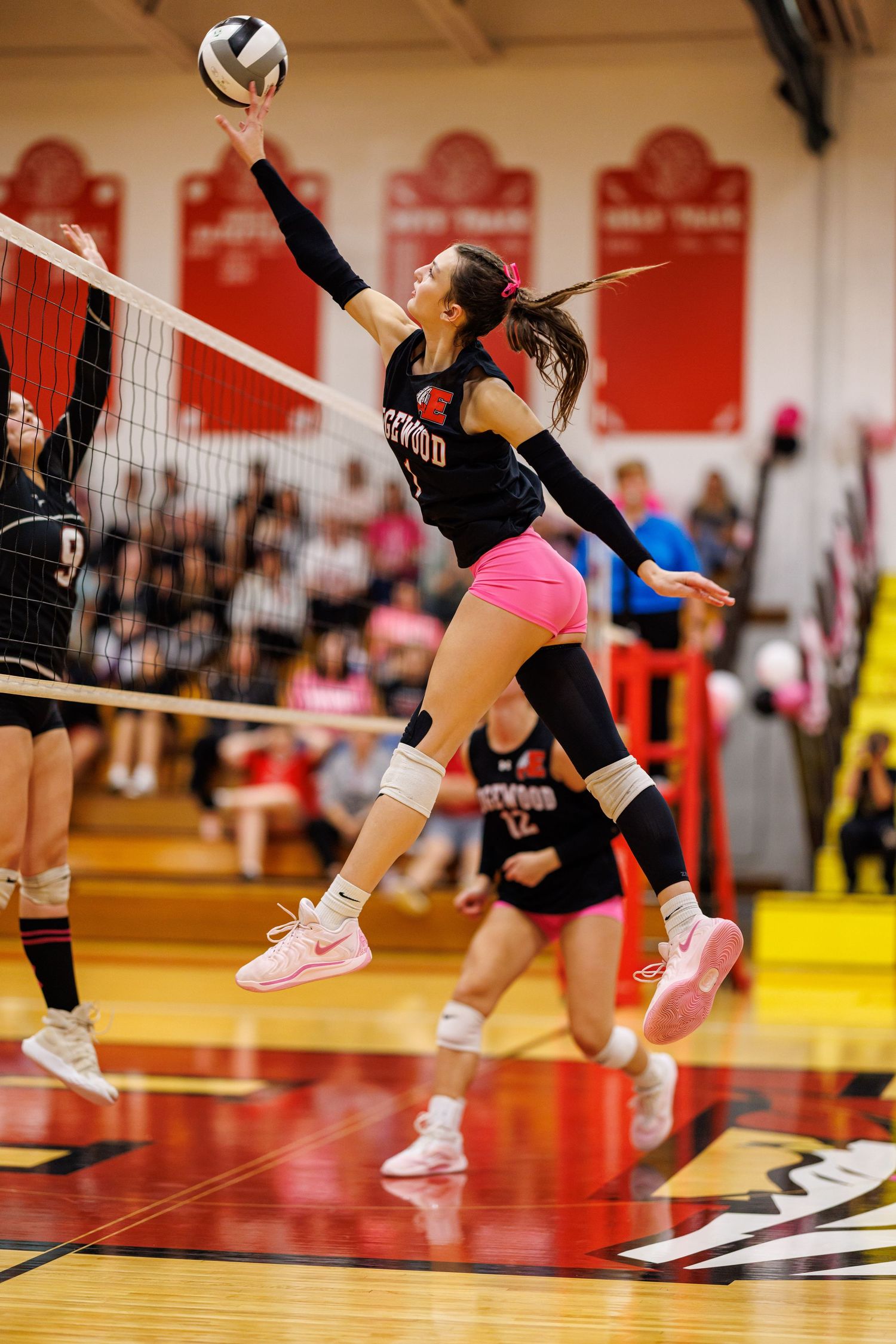 Female volleyball player in black and pink uniform leaps to spike the ball over the net during a game.