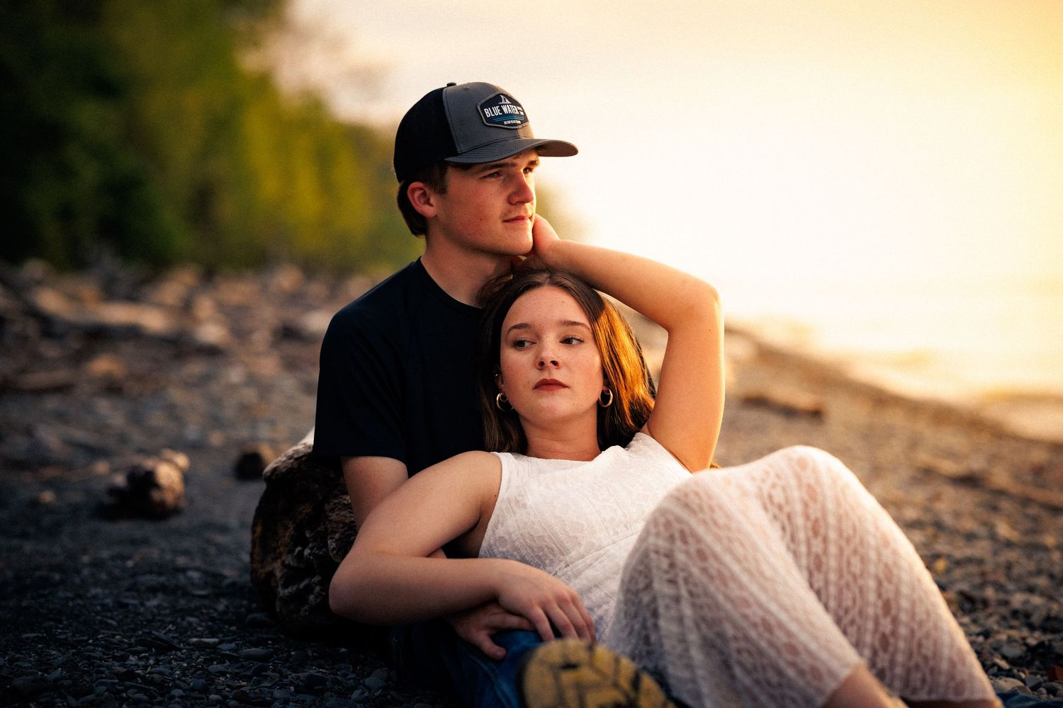 A romantic couple sitting together on a rocky beach at golden sunset, woman leaning against mans chest.