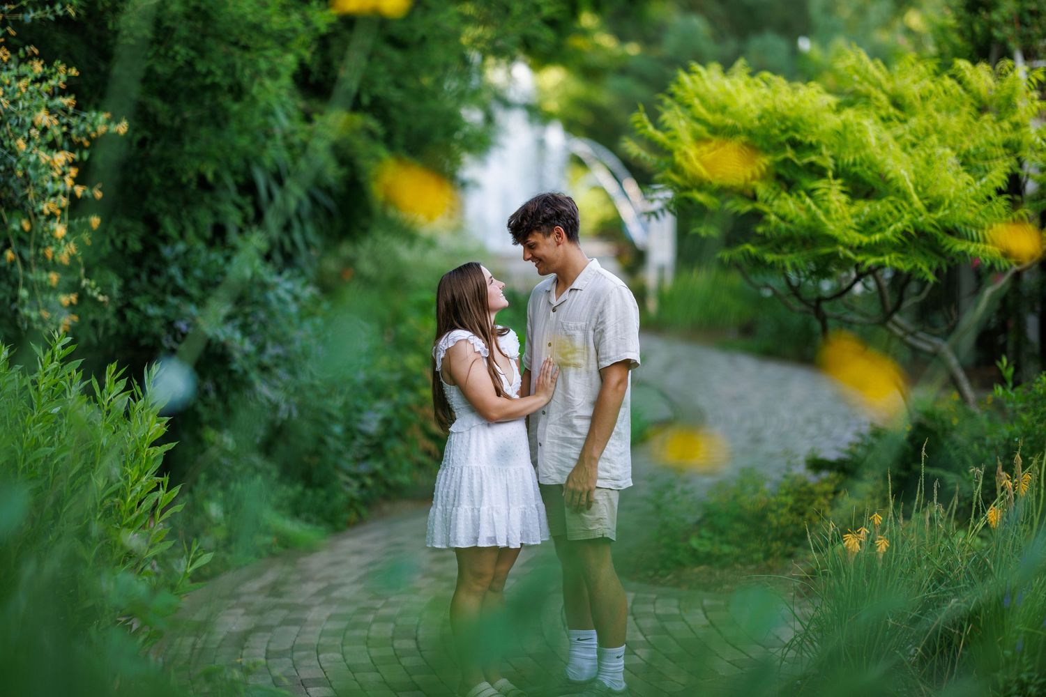 A young couple standing close together on a stone garden path surrounded by lush green foliage.