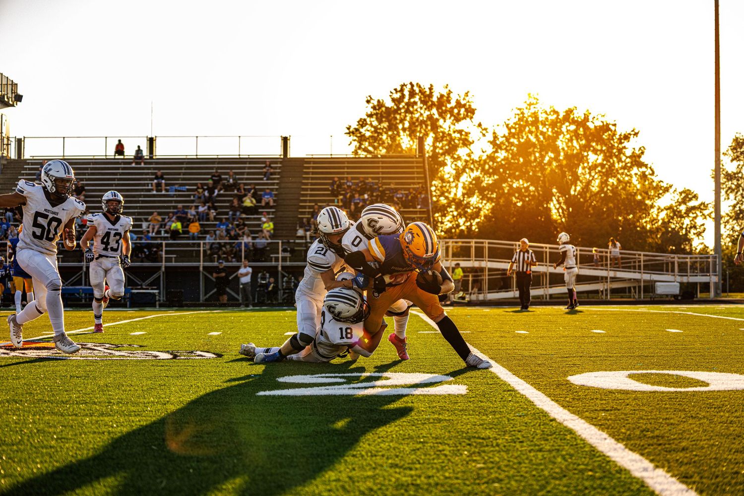Football players tackle a ball carrier at sunset on a green field with bleachers in the background.