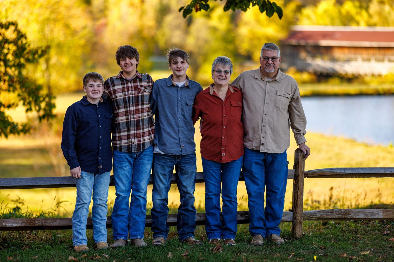 A family of five smiling together outdoors in fall, standing by a wooden fence with golden autumn trees in background.