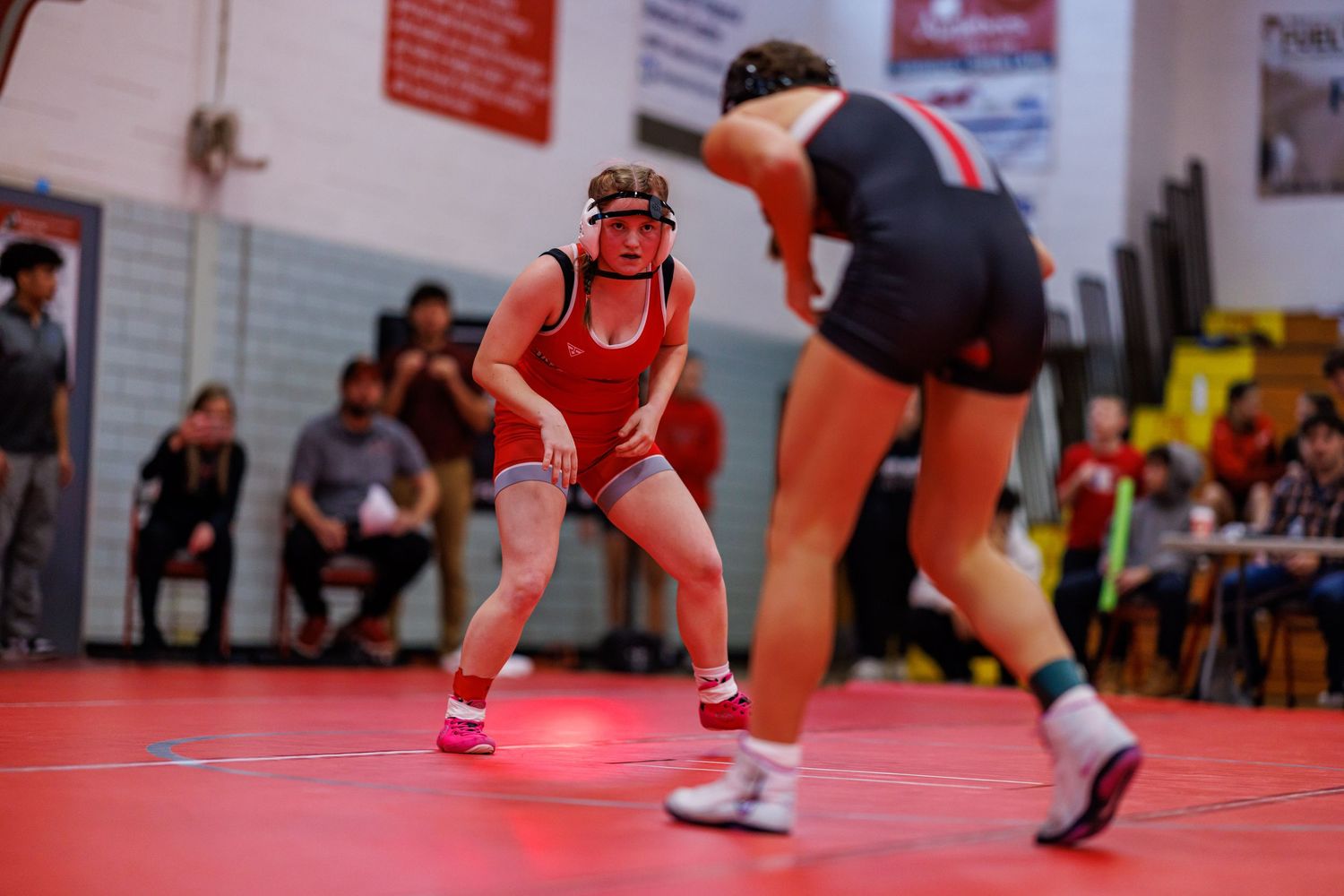 Two wrestlers in red uniforms face off on a red mat during a competitive wrestling match in a gymnasium.