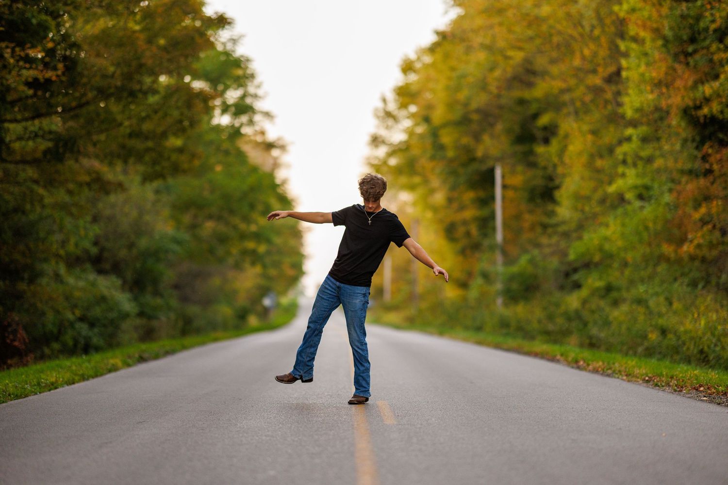 Teen boy in black shirt and jeans balancing on one foot on a scenic tree-lined road.