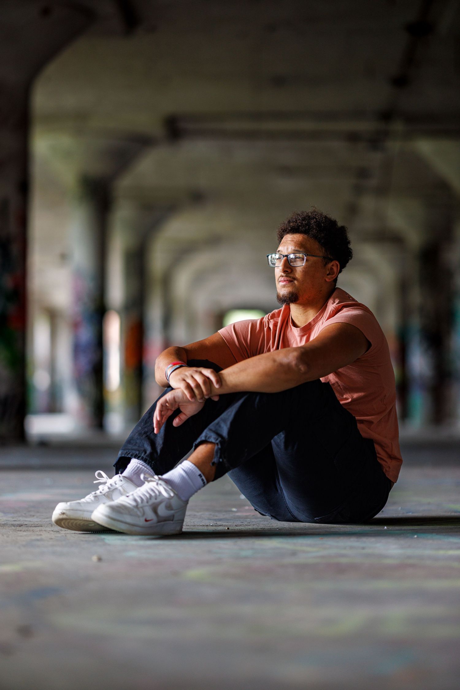 Young man in salmon shirt and white sneakers sitting on ground in graffiti-covered urban tunnel space.