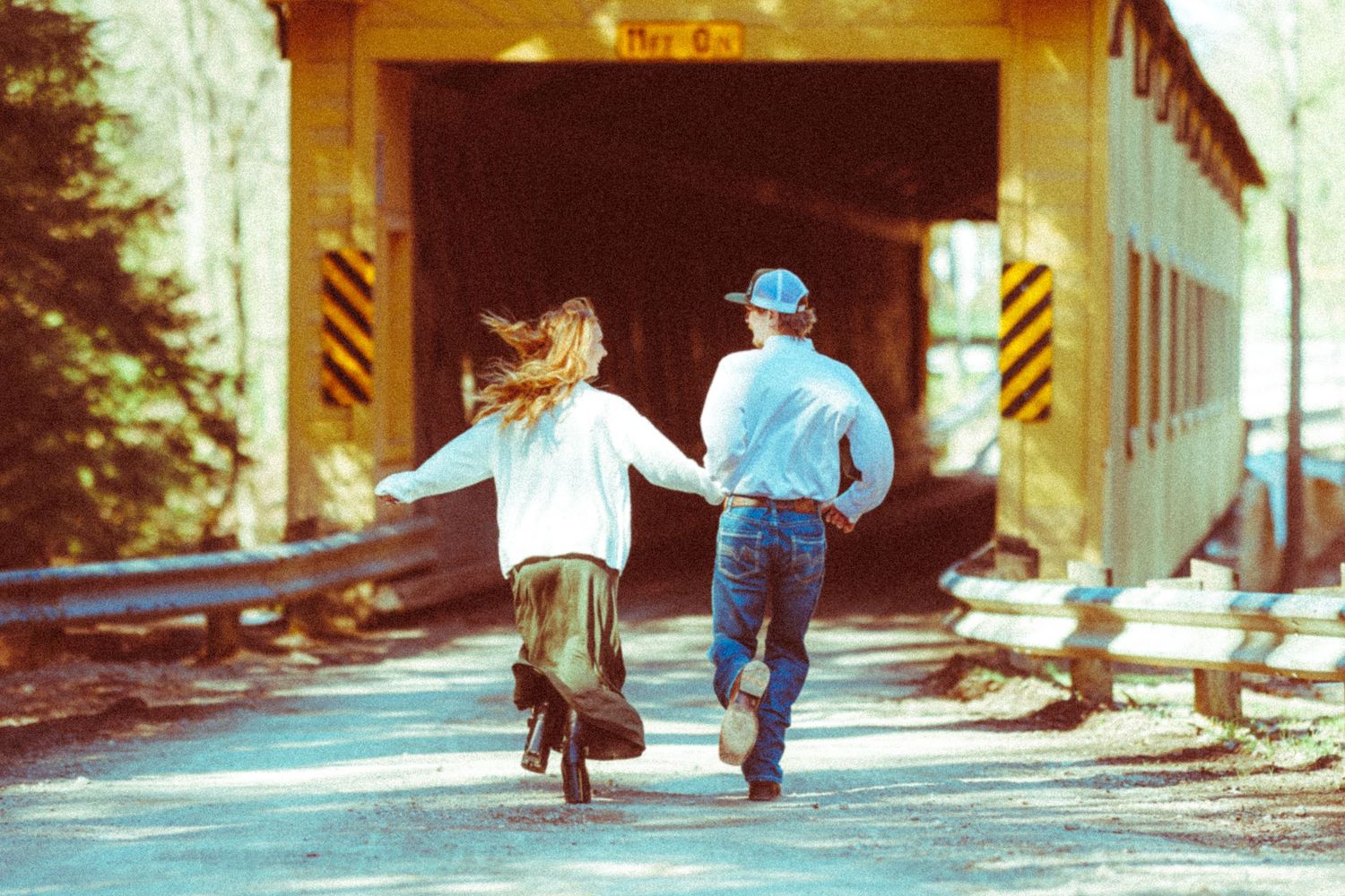 Couple runs hand-in-hand toward a yellow covered bridge on a sunny day.