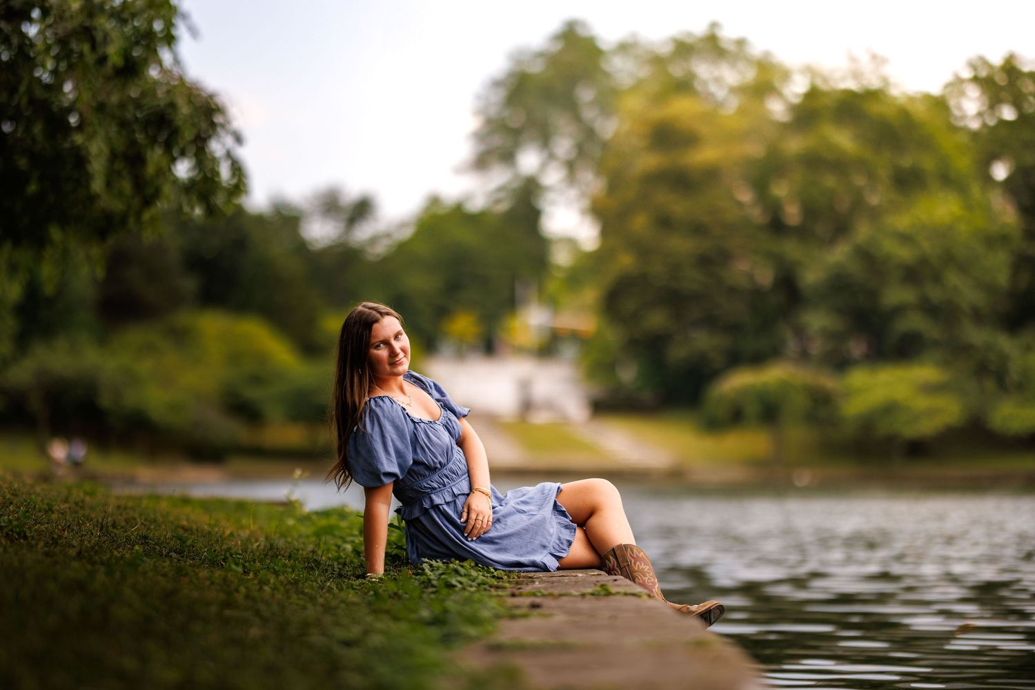 Woman in blue dress sitting by a tranquil park lake during golden hour.
